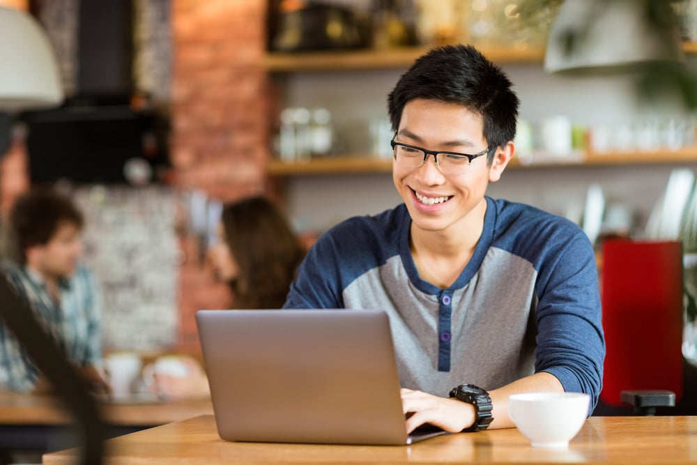 Happy cheerful young asian male in glasses smiling and using laptop in cafe to take part in an online community, representing online community engagement ideas