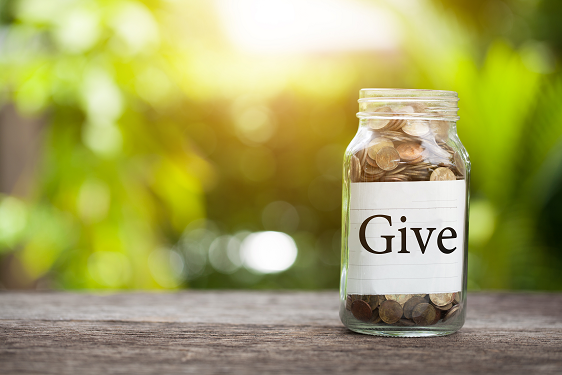 A jar full of coins on a table in front of a blurred green background with a label saying "Give," representing charity market research 
